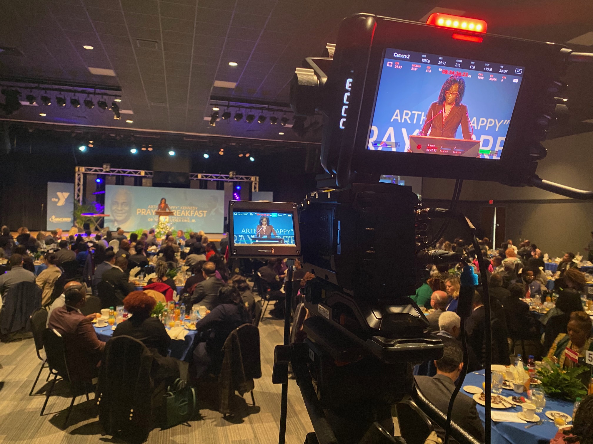 A broadcast camera capturing a speaker on stage during a full prayer breakfast event, with attendees seated at banquet tables